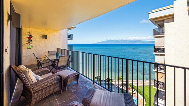 A balcony with wicker furniture, a table, and chairs overlooking a coastal view.