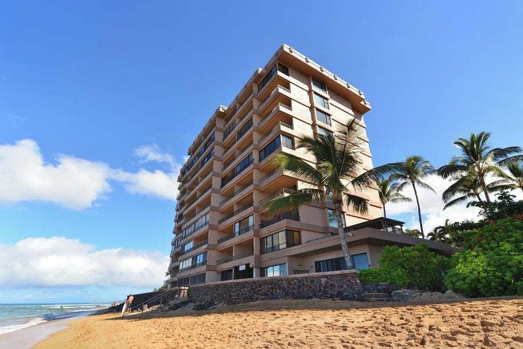 A multi-story hotel with balconies, located on a beach with palm trees and a clear sky.