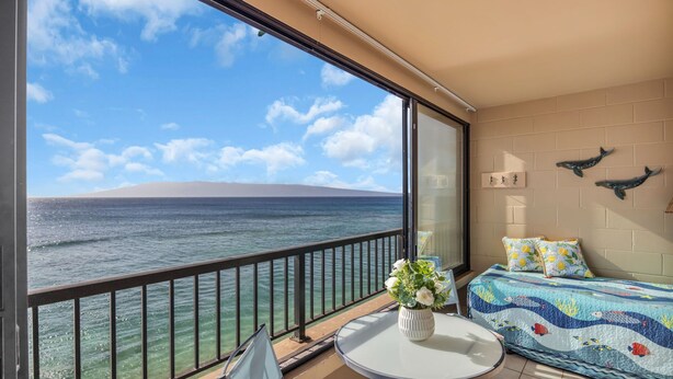 A balcony with a view of the ocean, a bed with a colorful quilt, and a small table with a vase of flowers. - MK404 - Sleep at the Ocean and Watch Sunsets 3-Bed Suite (Lahaina)