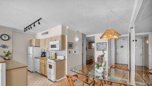 A modern kitchen with white appliances, a dining area with glass tables and wicker chairs, and a visible clock on the wall. - MK404 - Sleep at the Ocean and Watch Sunsets 3-Bed Suite (Lahaina)