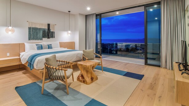 A modern bedroom with a large bed, a view of the city at dusk, and a balcony with a glass door.