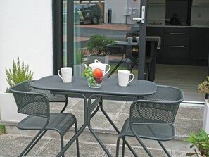 Een tafel met twee stoelen, een witte theepot en een schaal fruit met druiven en een appel.