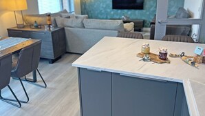 Moderni keittiö, jossa on keskeinen saareke, ruokailutila pöytineen ja tuoleineen, sohva sekä seinälle asennettu televisio.