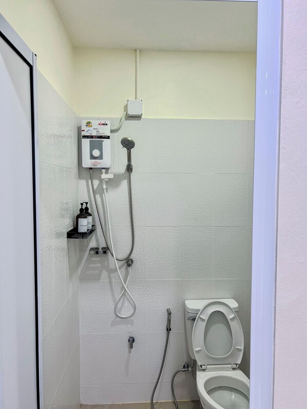Une petite salle de bain carrelée, équipée de toilettes, d’une douche et d’un distributeur de savon placé sur une étagère.