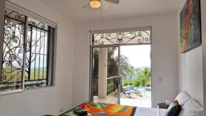 Une chambre avec un lit, un ventilateur de plafond, une fenêtre ornée de barreaux en fer décoratifs, et une vue sur une piscine et la verdure environnante.
