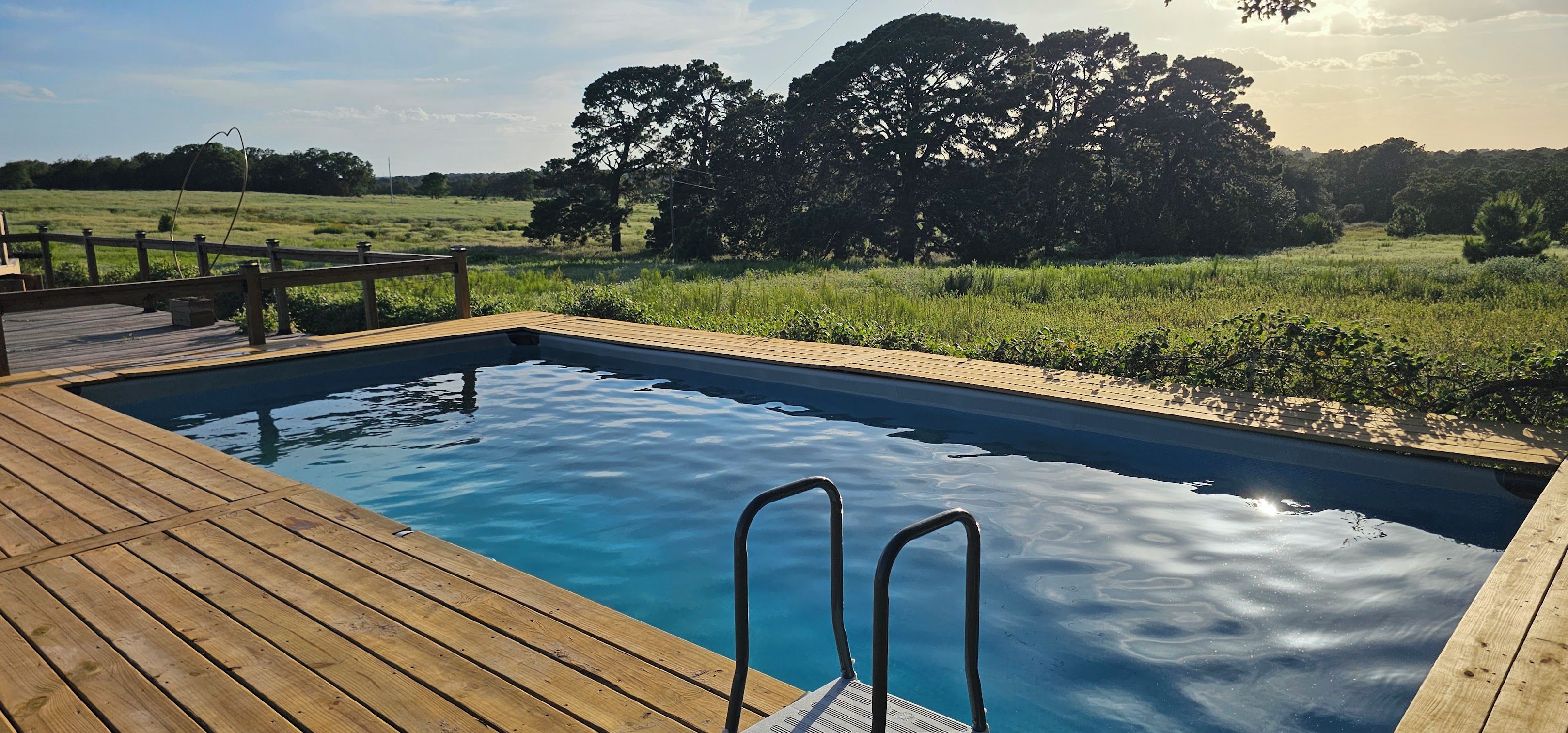 Ein rechteckiger Pool mit Holzterrasse und Metallleiter, umgeben von einer Rasenfläche und Bäumen.