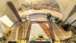 A spacious tent with a wooden floor, a bed with pillows, a green chair, and a dining area with a table and chairs. - Luxurious Stargazer Dome Retreat in Stunning Nature w/Hot Tub and Sauna! (Smithville)