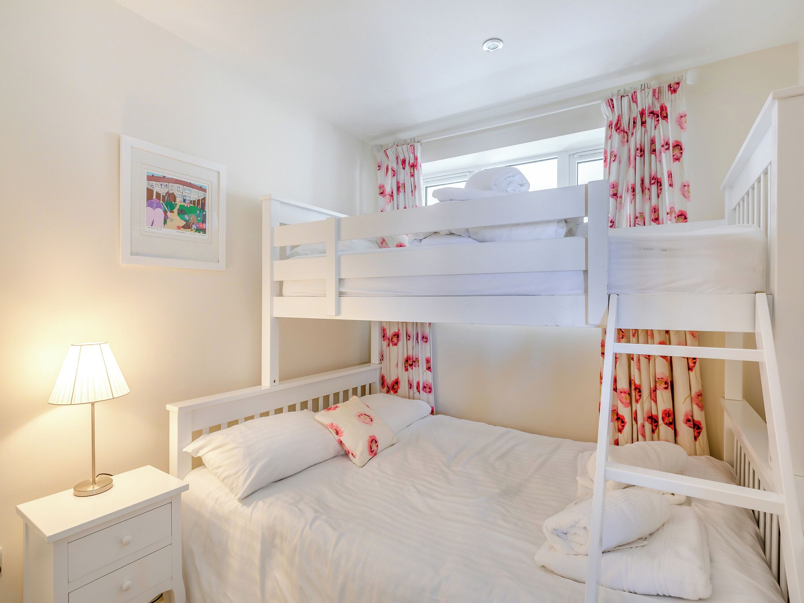 Habitación con literas, ropa de cama blanca, cortinas florales, una lámpara de noche y un cuadro en la pared.