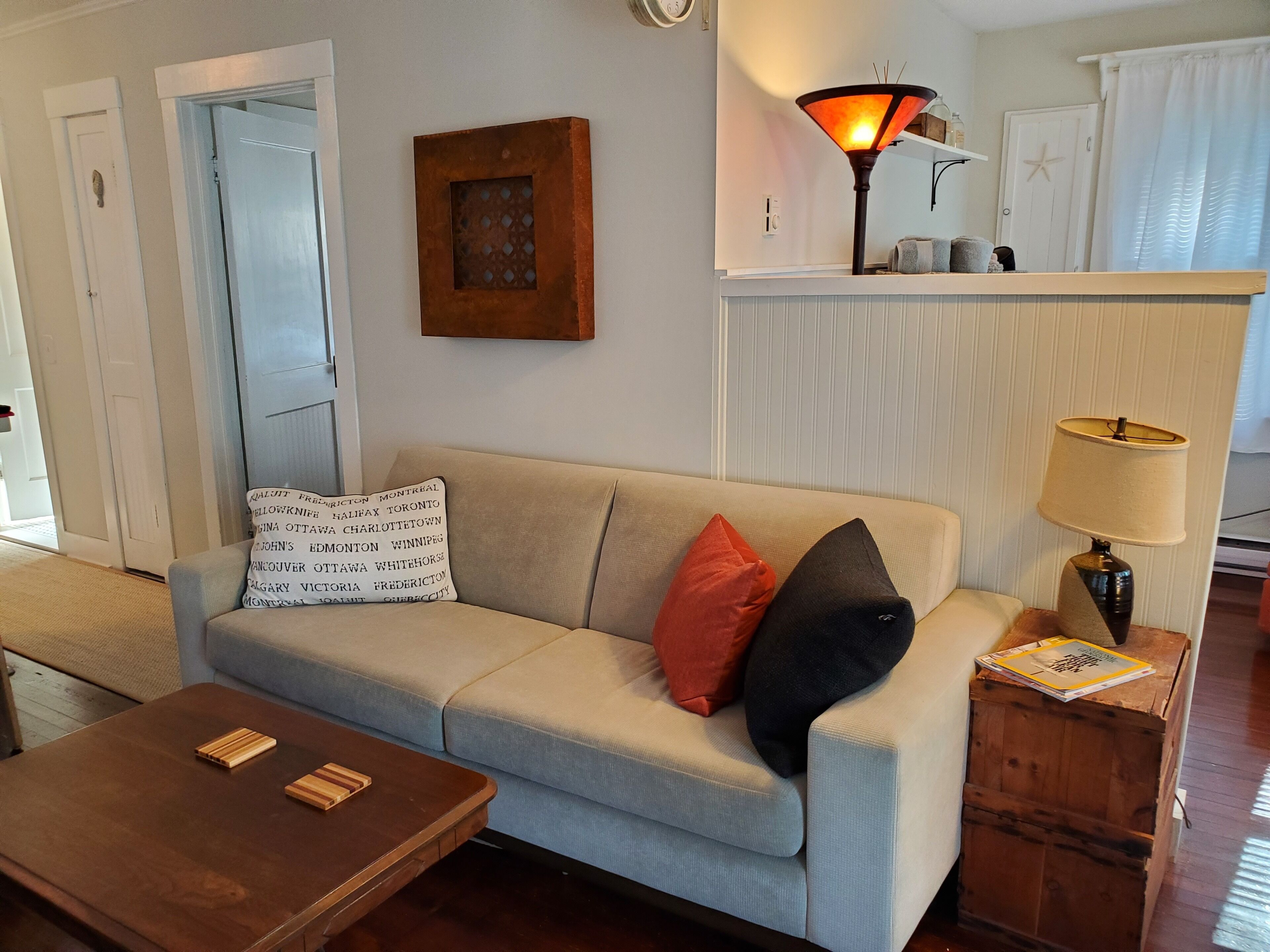 Un salon comprenant un canapé, deux coussins, une table basse en bois, une lampe et un petit meuble en bois.