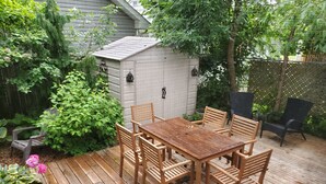 A wooden deck with a table and chairs, surrounded by lush greenery and a small shed.