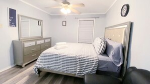 A bedroom with a bed, a dresser, a ceiling fan, a clock, and a window with blinds.