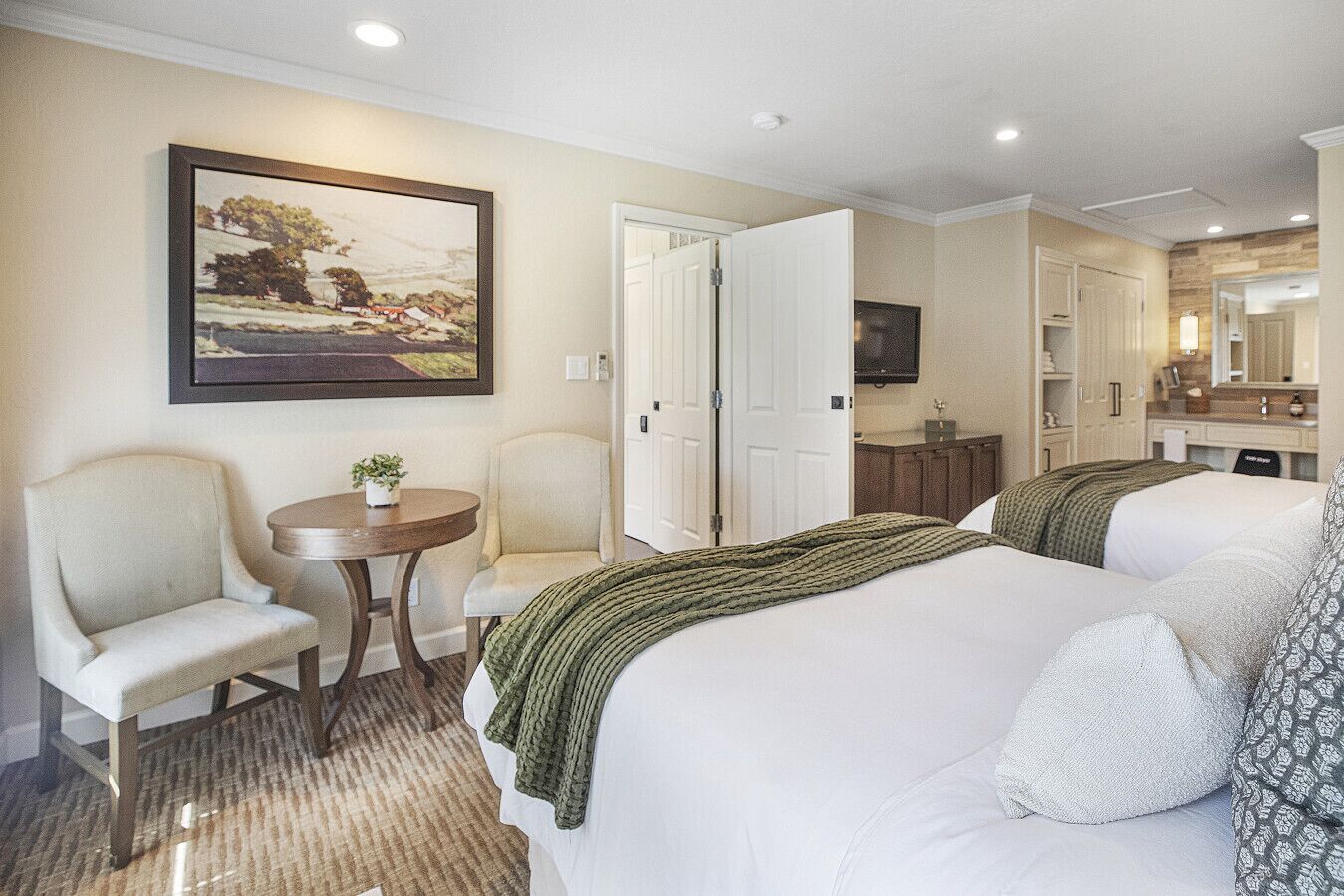 Una habitación de hotel con dos camas, una mesa redonda, una silla, un cuadro y un espejo.
