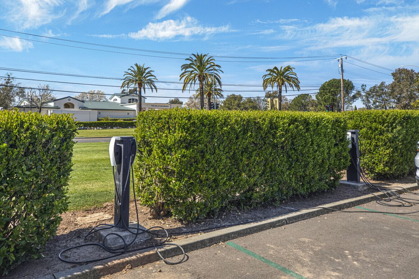 A parking area with electric vehicle charging stations, a hedge, and a building in the background.