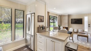 A modern kitchen with a stainless steel refrigerator, white cabinetry, and a large window with blinds.