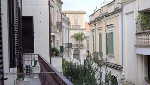 Una calle estrecha flanqueada por edificios, con un balcón adornado con plantas en macetas y un edificio que muestra un letrero que dice 'OSTA'.