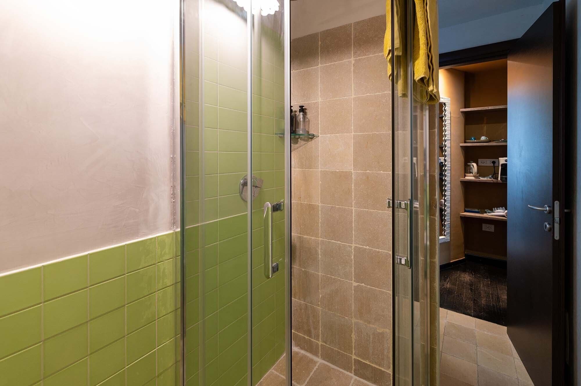 A bathroom with a glass shower enclosure, a green-tiled wall, and a beige-tiled floor.