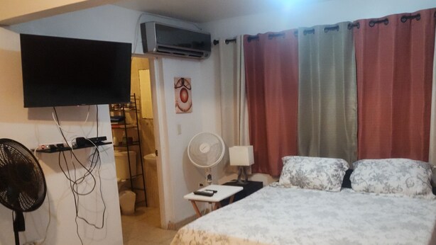 A hotel room with a bed, a TV mounted on the wall, a desk with a lamp, and a fan.