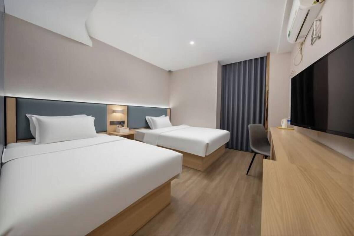 A hotel room with two beds, a desk, a chair, a flat-screen TV, and a window with curtains.