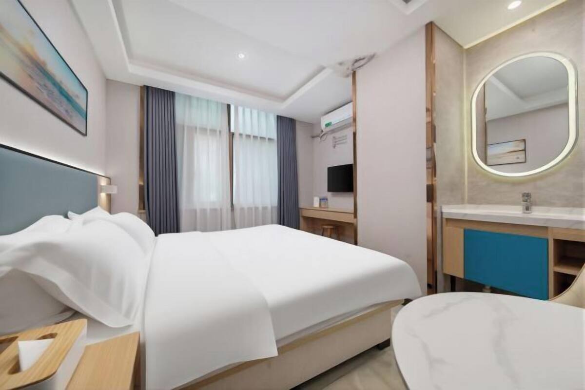 A hotel room with a bed, bedside table, a desk, a chair, a bathroom with a mirror, and a window with curtains.