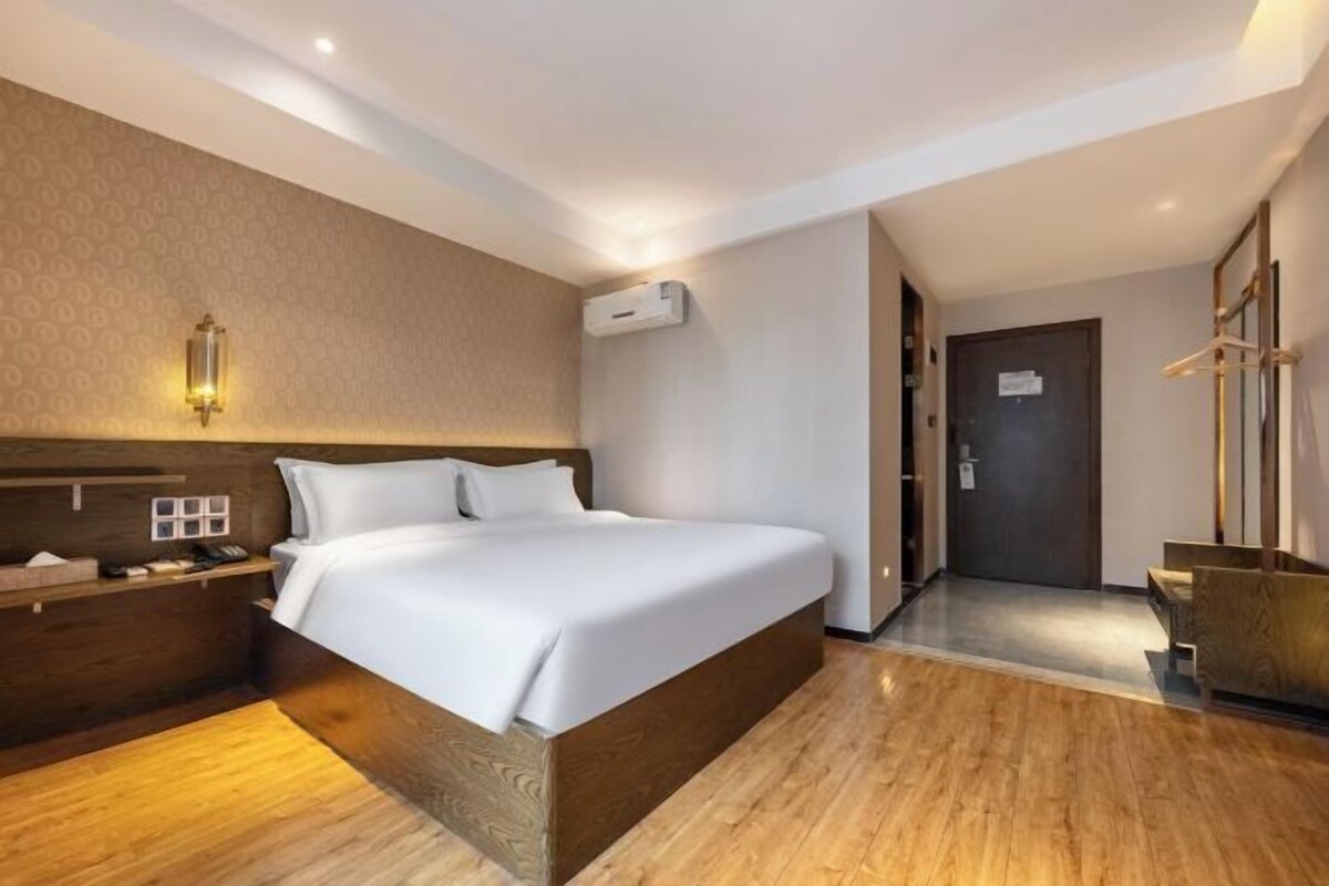 A hotel room with a wooden bed, a nightstand, a wall-mounted lamp, an air conditioning unit, and a door leading to another room.