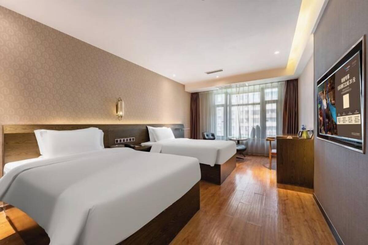 A hotel room with two beds, a desk, a chair, a window with curtains, and a wall-mounted light fixture.