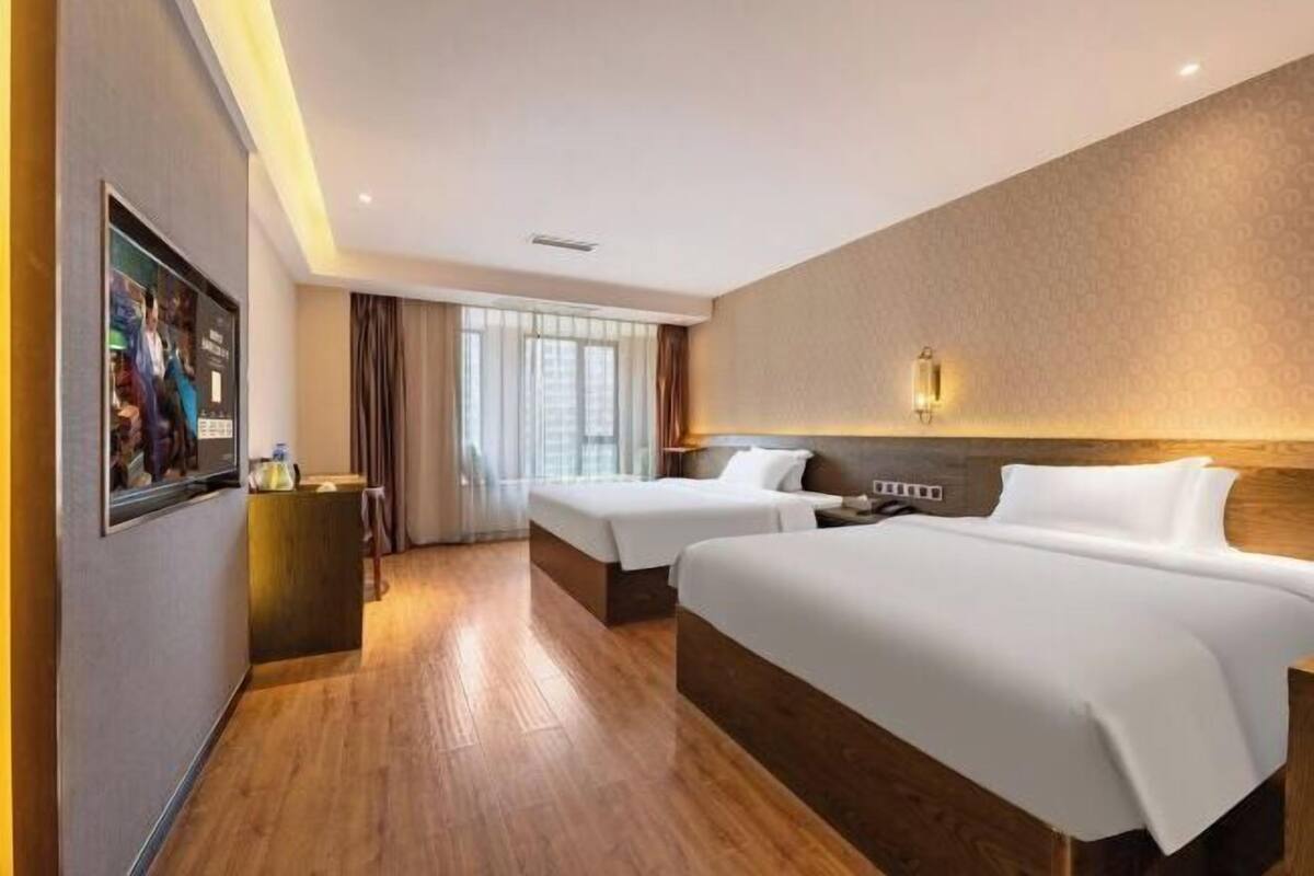 A hotel room with two beds, a wooden floor, a large window with curtains, and a wall-mounted TV.