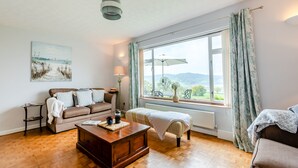 A cozy living room with a wooden coffee table, a sofa, and a view of the outdoors through a large window. - 3 bedroom accommodation in Membury (Membury)