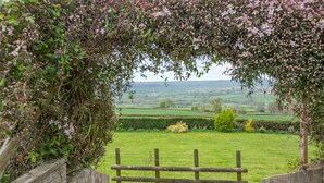 A stone pathway leading through a wooden gate into a lush green field with a view of rolling hills in the distance. - 3 bedroom accommodation in Membury (Membury)