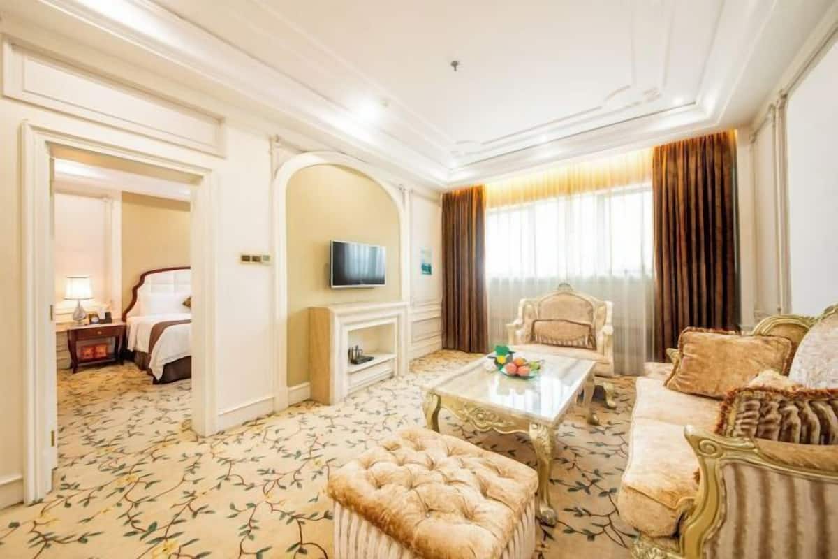 A spacious living room with a sofa, ottoman, and a coffee table. There is a television mounted on the wall and a window with curtains.