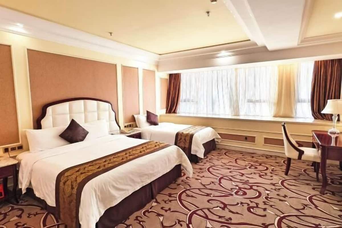 A hotel room with two beds, a large window with curtains, a patterned carpet, and a desk with a chair.