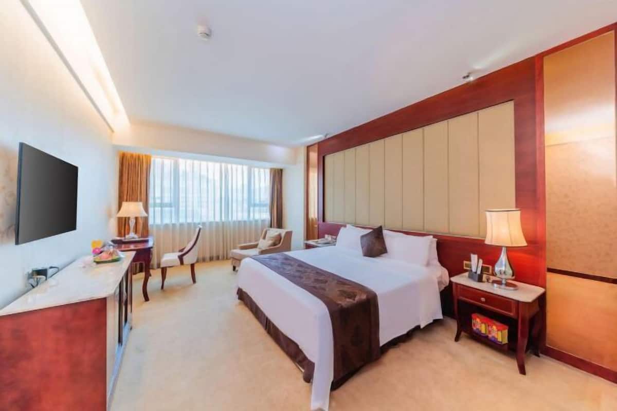 A hotel room with a bed, bedside table, lamp, chair, desk, TV, and a window with curtains.