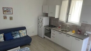 Une cuisine compacte avec des meubles blancs, un four encastré et un évier. On y trouve une petite table à manger recouverte d’une nappe à motifs.