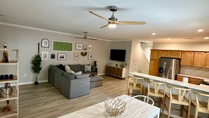 A modern living room with a grey sofa, a dining table with chairs, a ceiling fan, and a kitchen area with wooden cabinets and a refrigerator. - 4br/3ba + pool, close to beach. Pet friendly, WiFi, Parking, Quiet neighborhood (Rehoboth Beach)