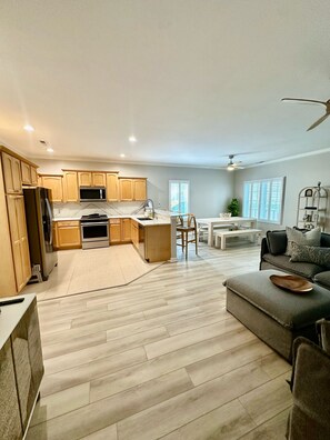 Une cuisine moderne avec des meubles en bois, un réfrigérateur et un four à micro-ondes. On y trouve un coin repas avec une table et des chaises blanches. Le salon est équipé d’un canapé gris et d’un ventilateur de plafond.