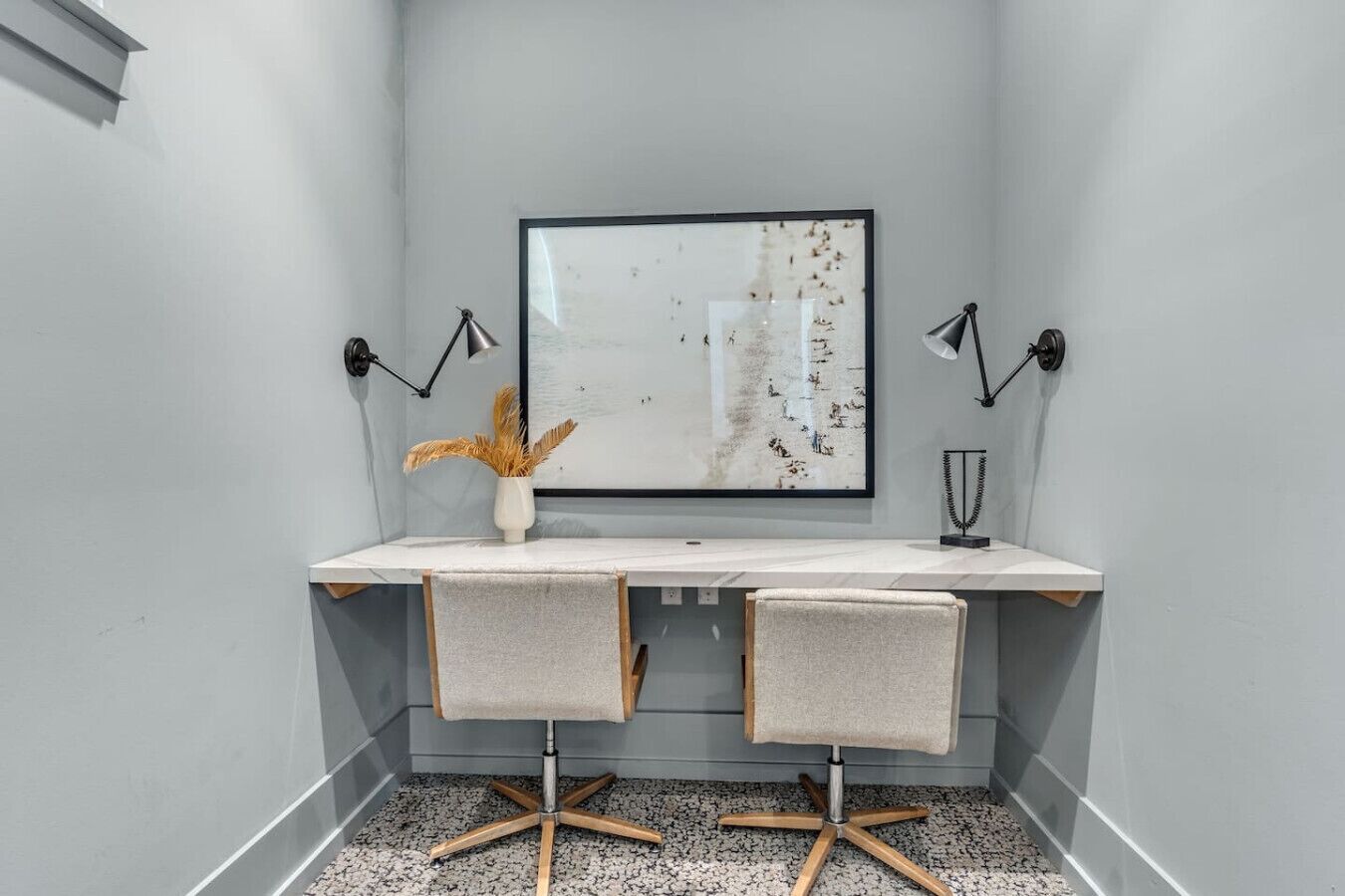 A modern office space with a white desk, two chairs, a large abstract painting, and two wall-mounted lamps.