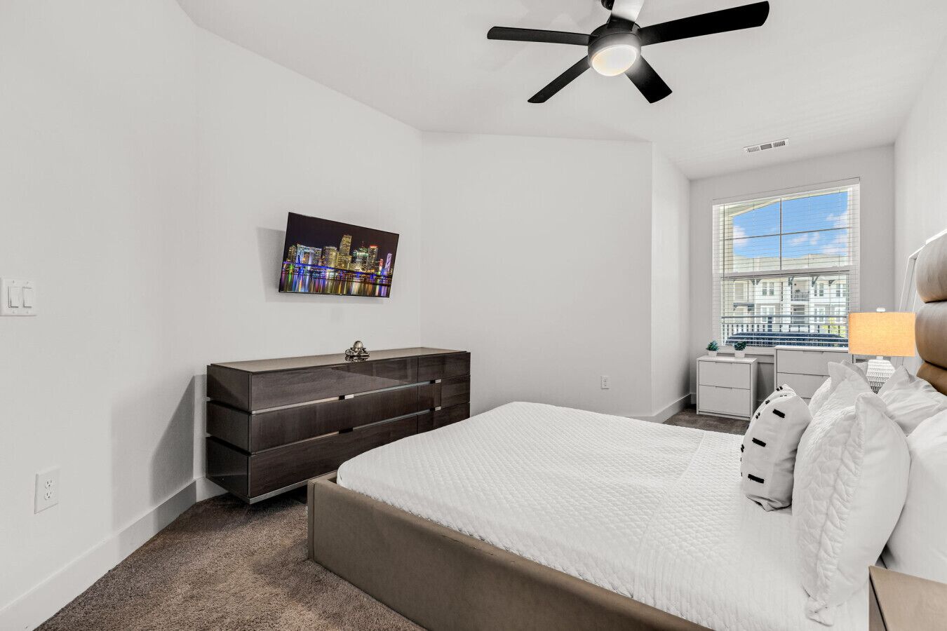 Une chambre à coucher comprenant un lit, une commode, un ventilateur de plafond, une fenêtre avec vue et un tableau représentant un paysage urbain.