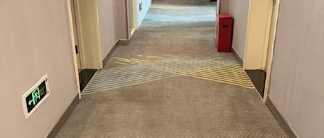 A carpeted hallway with directional arrows on the floor, a fire alarm panel, and a red trash bin.