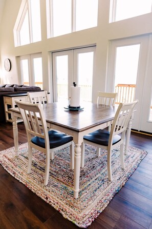 Un coin repas avec une table en bois et des chaises blanches, un tapis coloré et de grandes fenêtres.