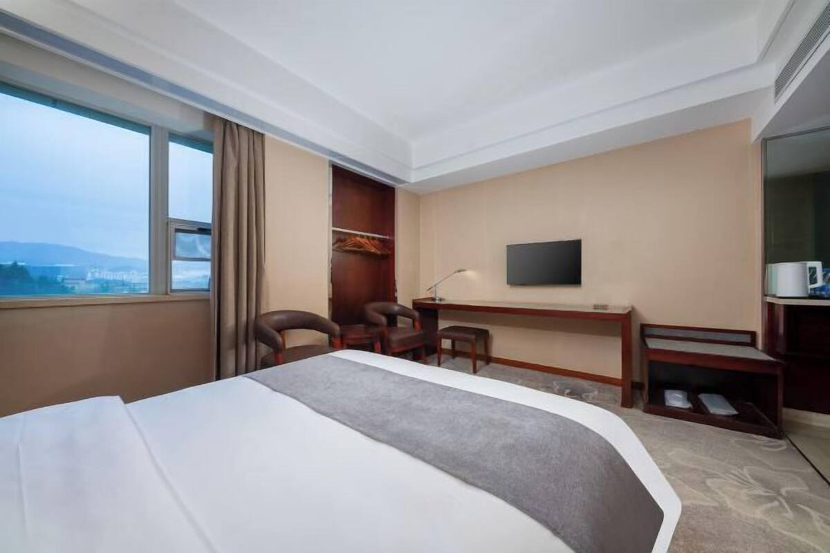 A hotel room with a bed, desk, chair, TV, and a view of the city.