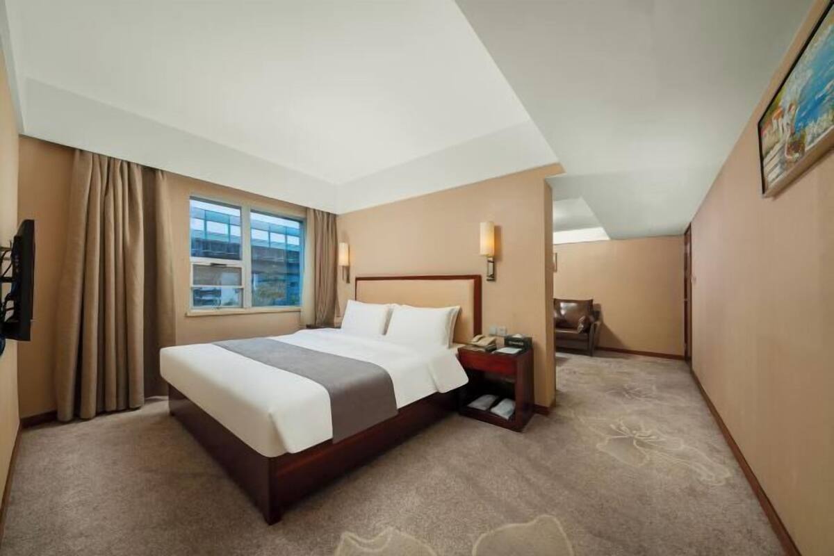 A hotel room with a large bed, a bedside table with a lamp, a window with curtains, and a painting on the wall.