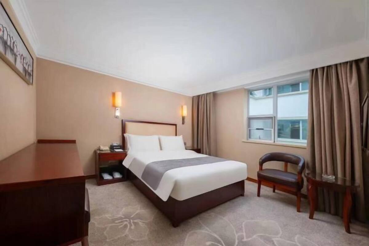 A hotel room with a bed, bedside table, lamp, chair, and window with curtains.