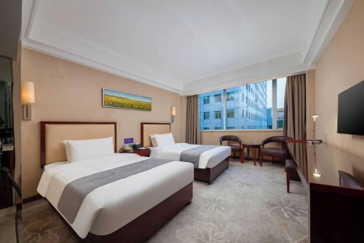 A hotel room with two beds, a desk, a chair, a TV, and a large window.