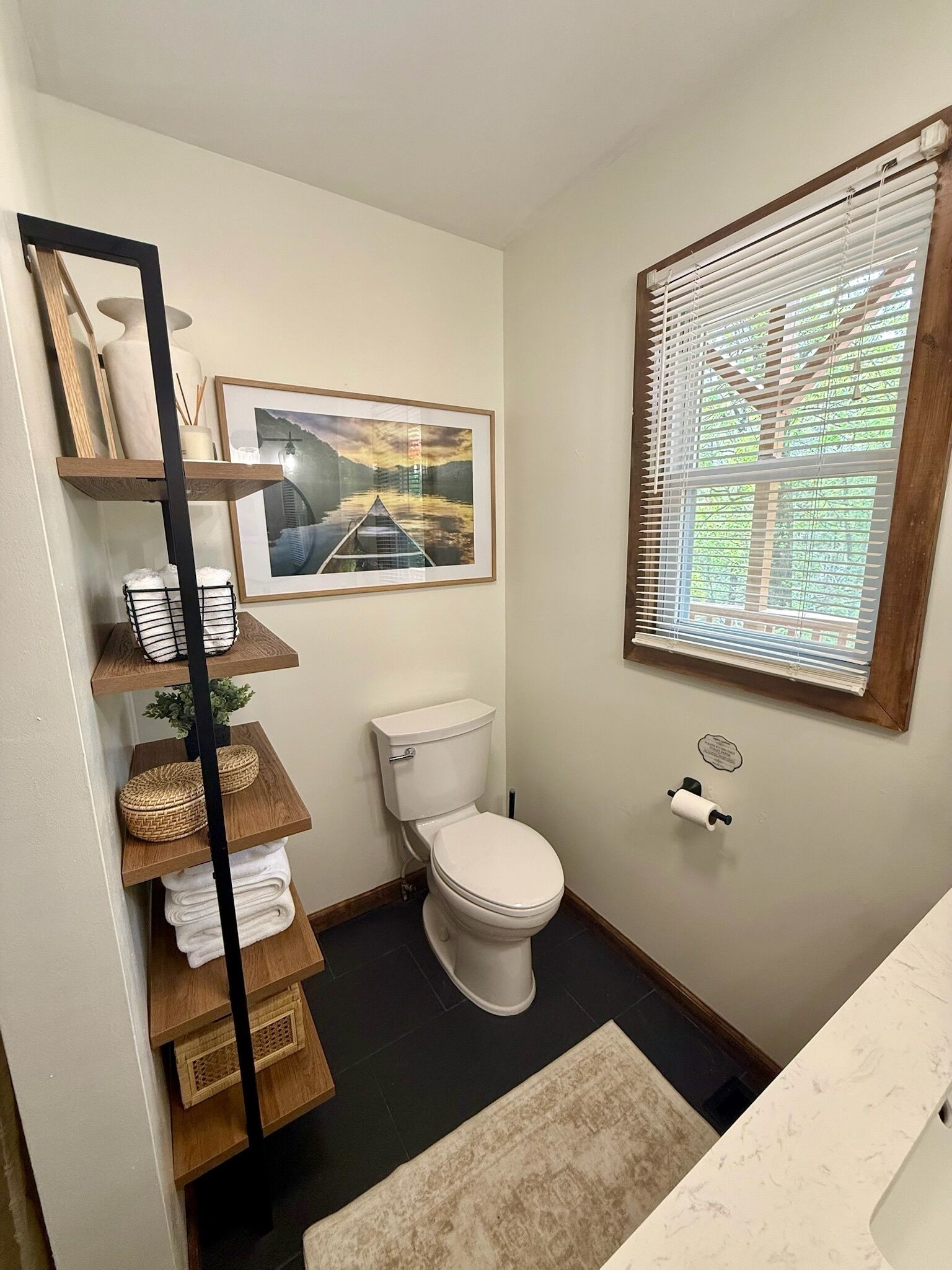 A bathroom with a toilet, a framed picture on the wall, a window with blinds, and a shelf with towels and baskets.