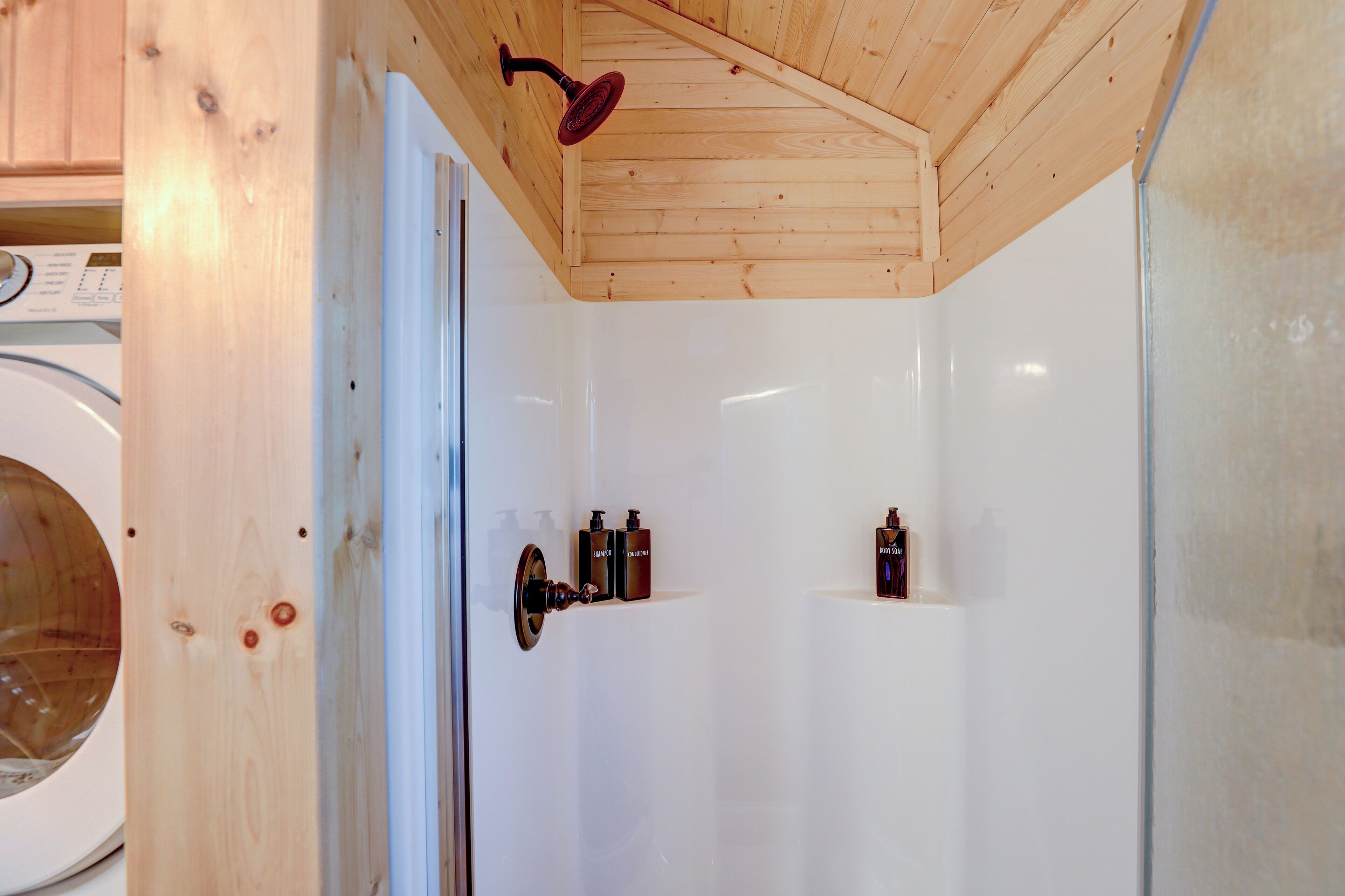 A small, enclosed shower area with wooden ceiling and white walls, featuring a built-in hairdryer and two bottles of shampoo.
