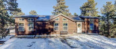A two-story wooden cabin with a bench in front, surrounded by snow and trees.