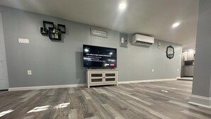 A spacious living room with a large flat-screen TV on a stand, grey walls, and a wooden floor. - Beach Block Cozy studio with WiFi and FREE PARKING!! (Atlantic City)