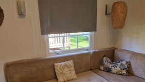 A cozy living room with a beige sofa, a small round table, and a window with a view of a wooden deck. - Mobile home number 480 in camping 4☆ (Châteauneuf-de-Galaure)