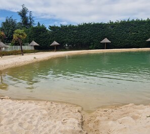 A sandy beach with a calm pool, surrounded by trees and thatched umbrellas.