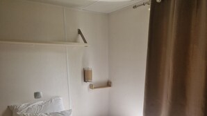 A single bed with white bedding, a bedside table, a wall-mounted light, and a window with brown curtains. - Mobile home number 480 in camping 4☆ (Châteauneuf-de-Galaure)