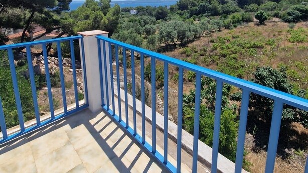 Un balcon doté d’une balustrade bleue, d’où l’on peut admirer un paysage arboré et une vue lointaine sur la mer.